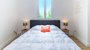 Ein ordentlich bezogenes Bett mit weißer Decke und rotem Kopfkissen, zwei Nachttischlampen, ein Fenster mit Blick und eine Wand mit motivierenden Sprüchen.