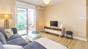 Ein modernes Wohnzimmer mit einer Couch, einem Glastisch, einem Fernseher auf einem Holzgestell und einem kleinen Hocker.