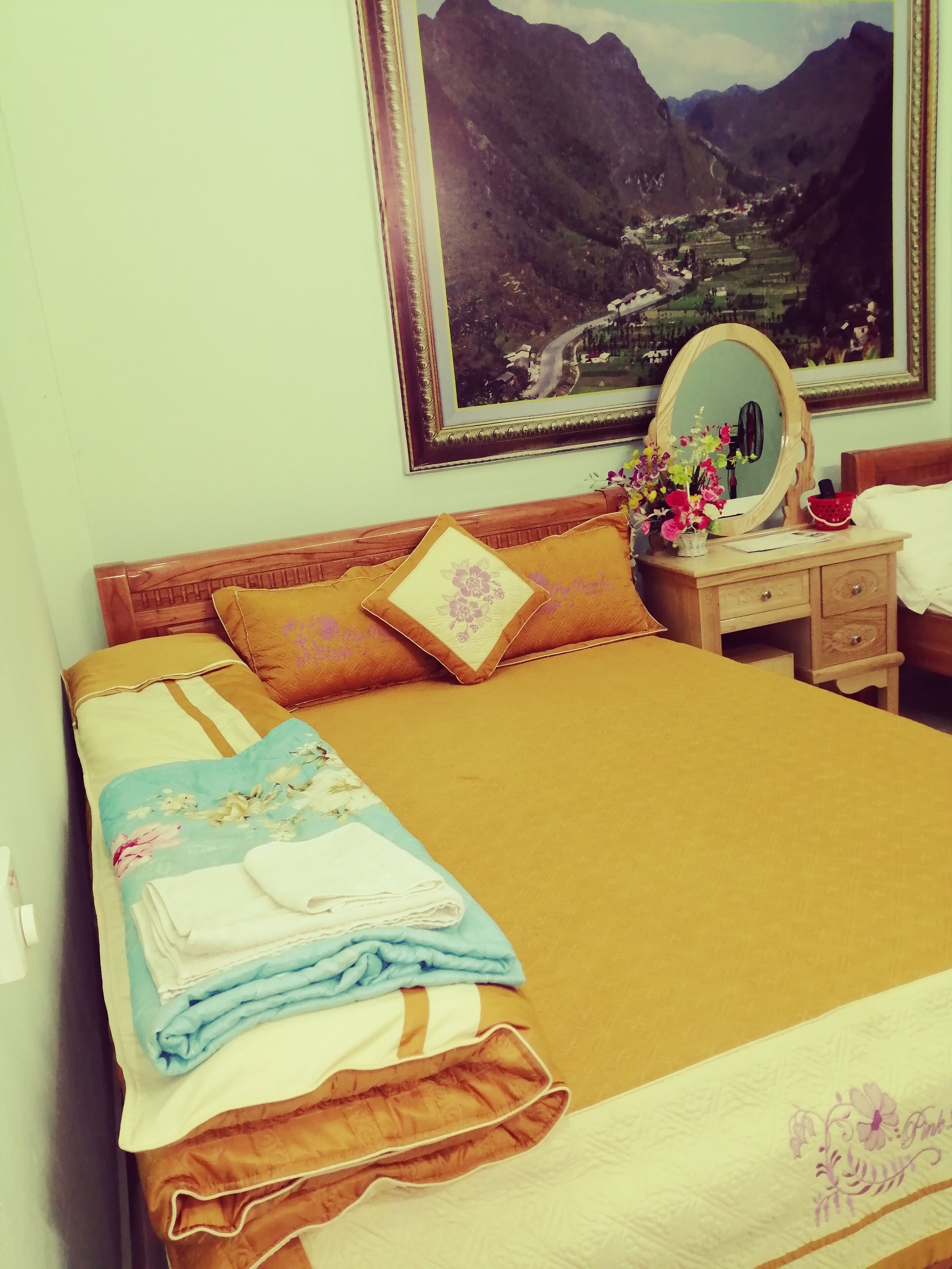 A neatly made bed with a floral bedspread, a wooden headboard, and a nightstand with a mirror and a vase of flowers.