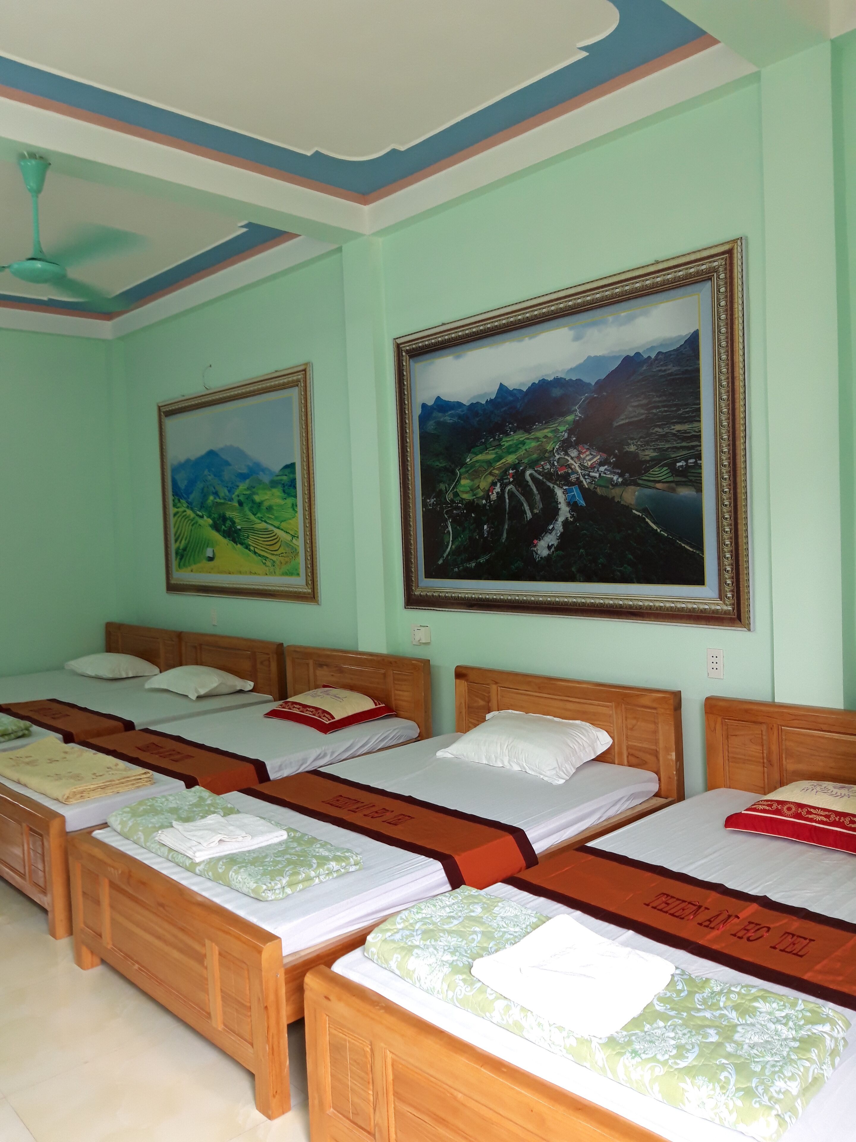 A hotel room with four wooden beds, each with a white mattress and a green and white patterned bedspread. There are two framed landscape paintings on the wall, one depicting a mountainous landscape and the other a river scene.