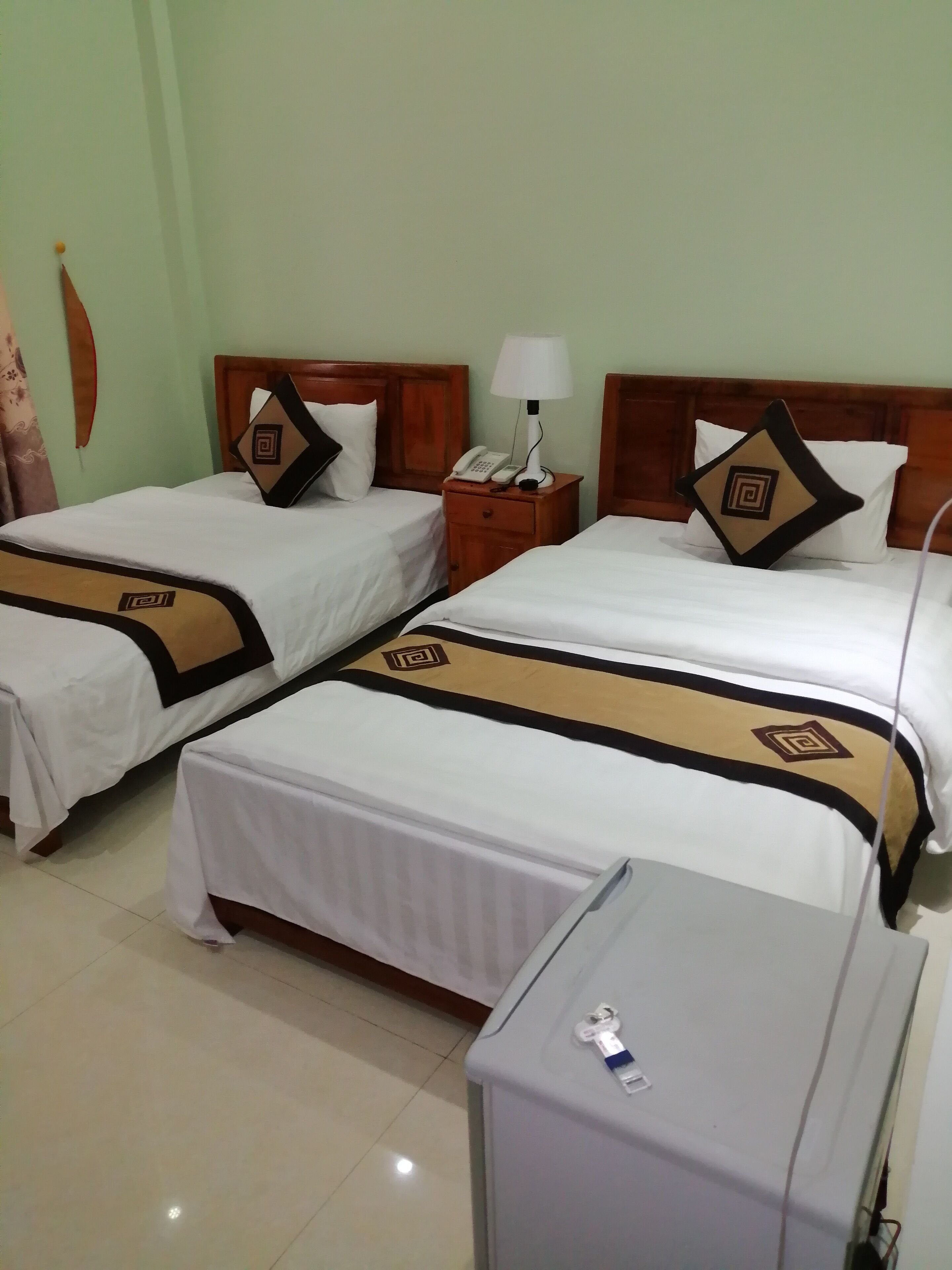 A hotel room with two beds, each with a wooden headboard, white bedding, and decorative pillows. There is a bedside table with a lamp and a telephone.