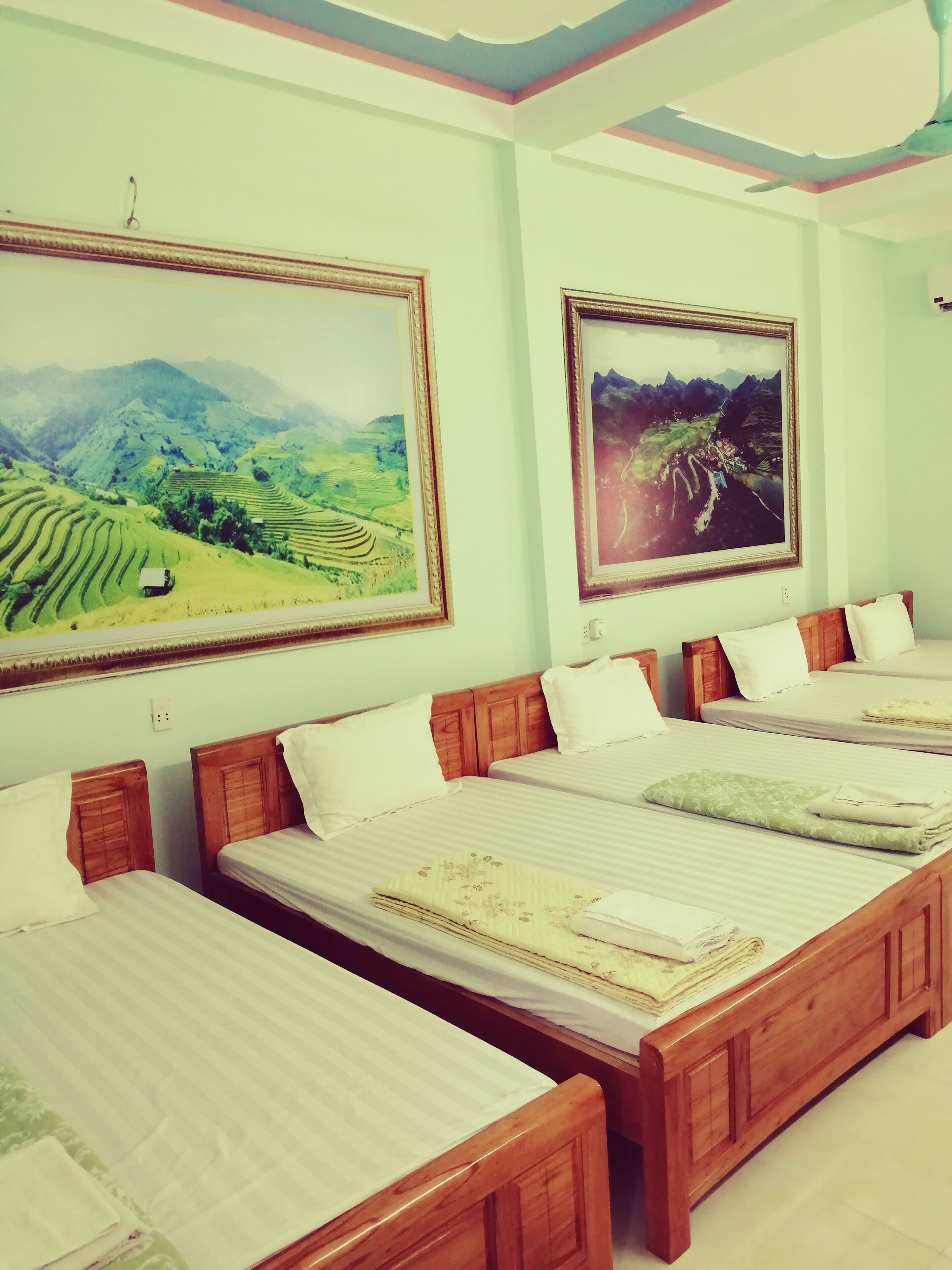 A hotel room with three beds, each with wooden headboards and white bedding. There are two large landscape paintings on the walls, one depicting a green valley and the other a mountainous landscape.