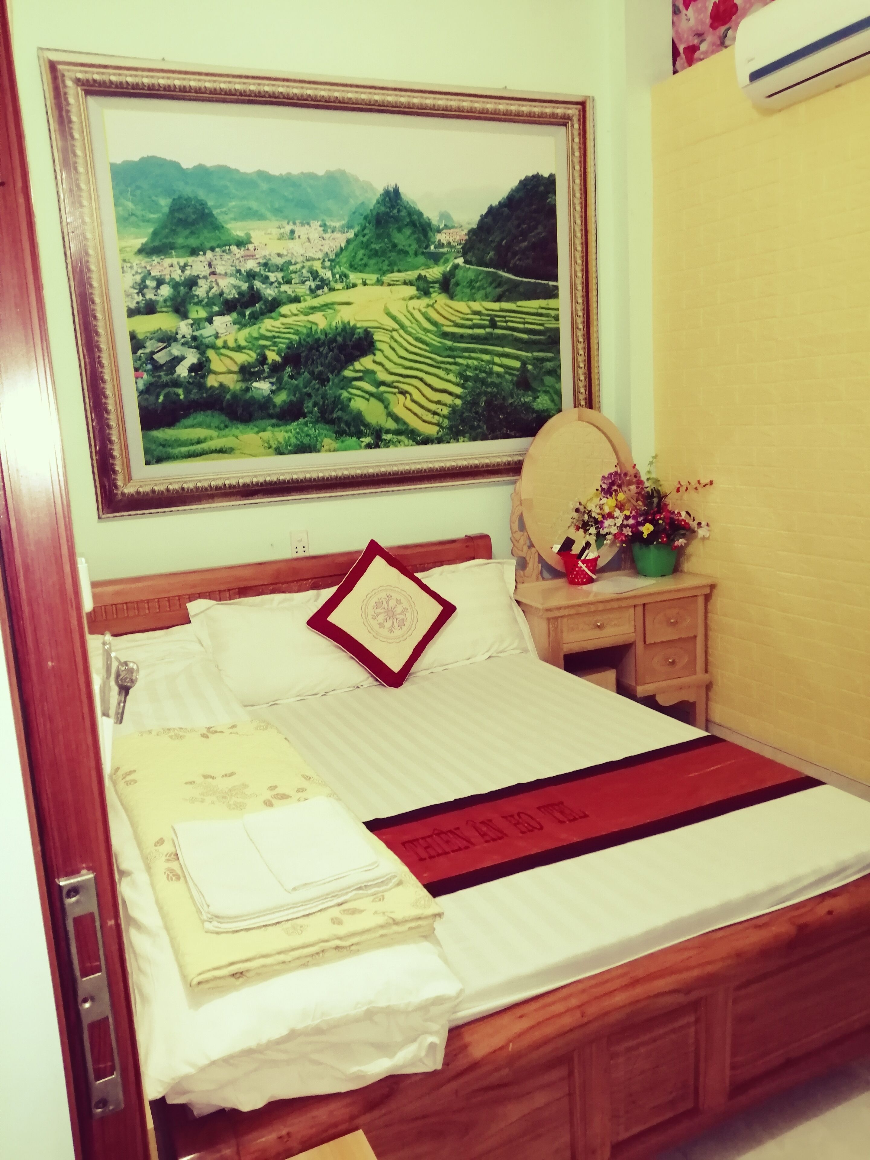 Ein ordentlich bezogenes Bett mit einer rot-schwarzen Tagesdecke, einem Kissen mit Blumenmuster und einem Rahmenbild mit einer Landschaft darüber.