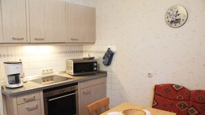 Una cocina compacta con microondas, estufa y cafetera. Un reloj en la pared, y un papel tapiz decorado cuelga sobre una mesa de madera con una cesta.