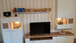 Una sala de estar moderna con un televisor de pantalla plana, muebles blancos y objetos decorativos en estanterías.
