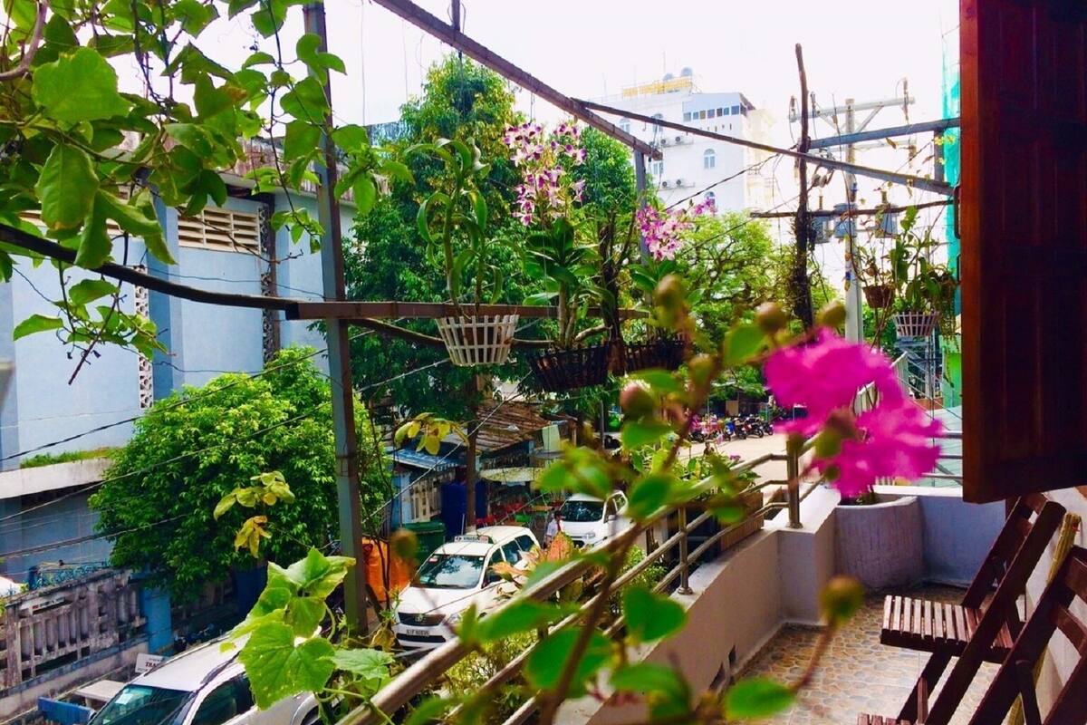 Een balkon met uitzicht op een drukke straat, groen en een roze bloem.