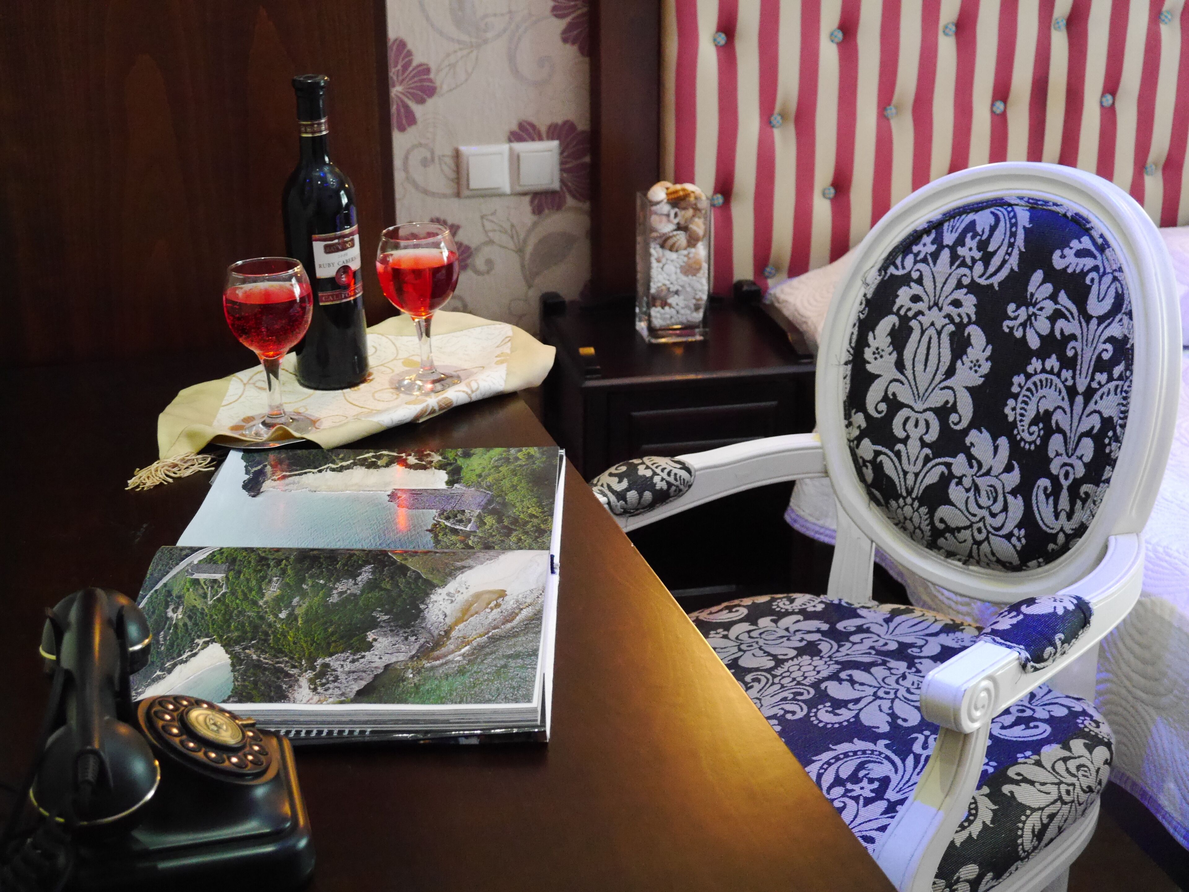Um quarto com uma mesa de madeira, uma cadeira com almofada estampada, uma garrafa de vinho, duas taças de vinho e um livro de capa com paisagem.