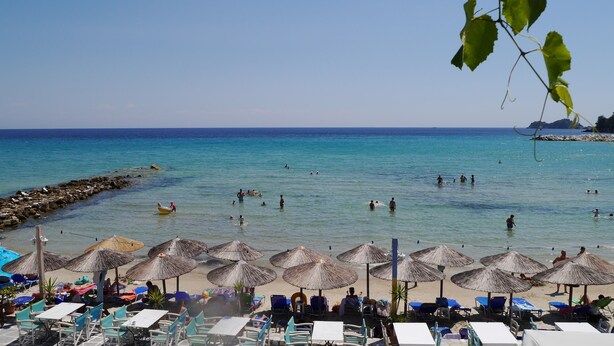 A beach with thatched umbrellas and lounge chairs, a rocky breakwater, and clear blue water. - Blue Sea Beach Hotel (Thasos)