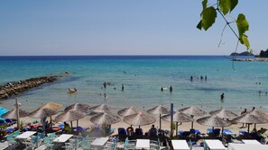 A beach with thatched umbrellas and lounge chairs, a rocky breakwater, and clear blue water. - Blue Sea Beach Hotel (Thasos)