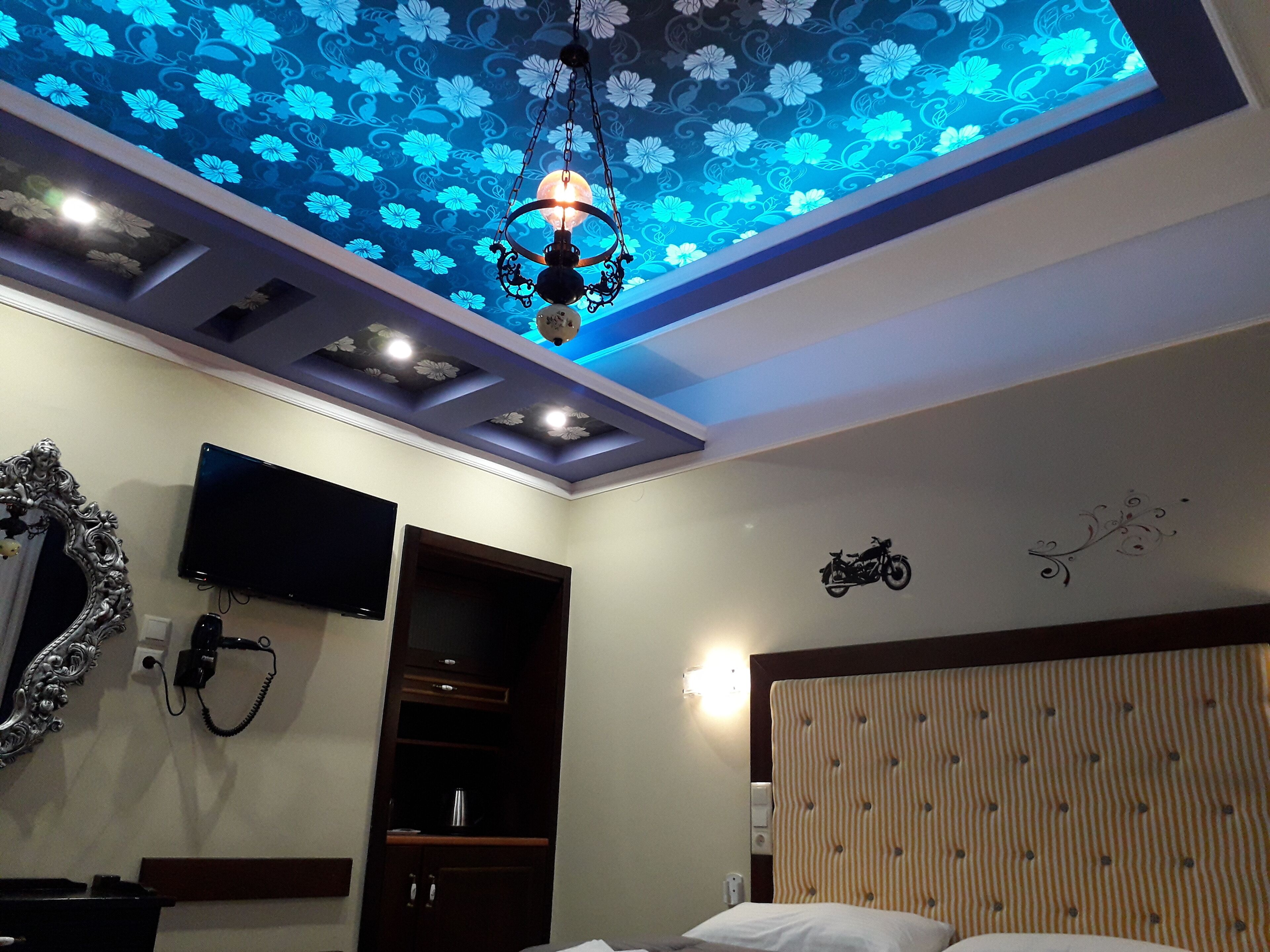 A hotel room with a ceiling with floral patterns, a mounted TV, a bedside table, a bed, and a wardrobe.