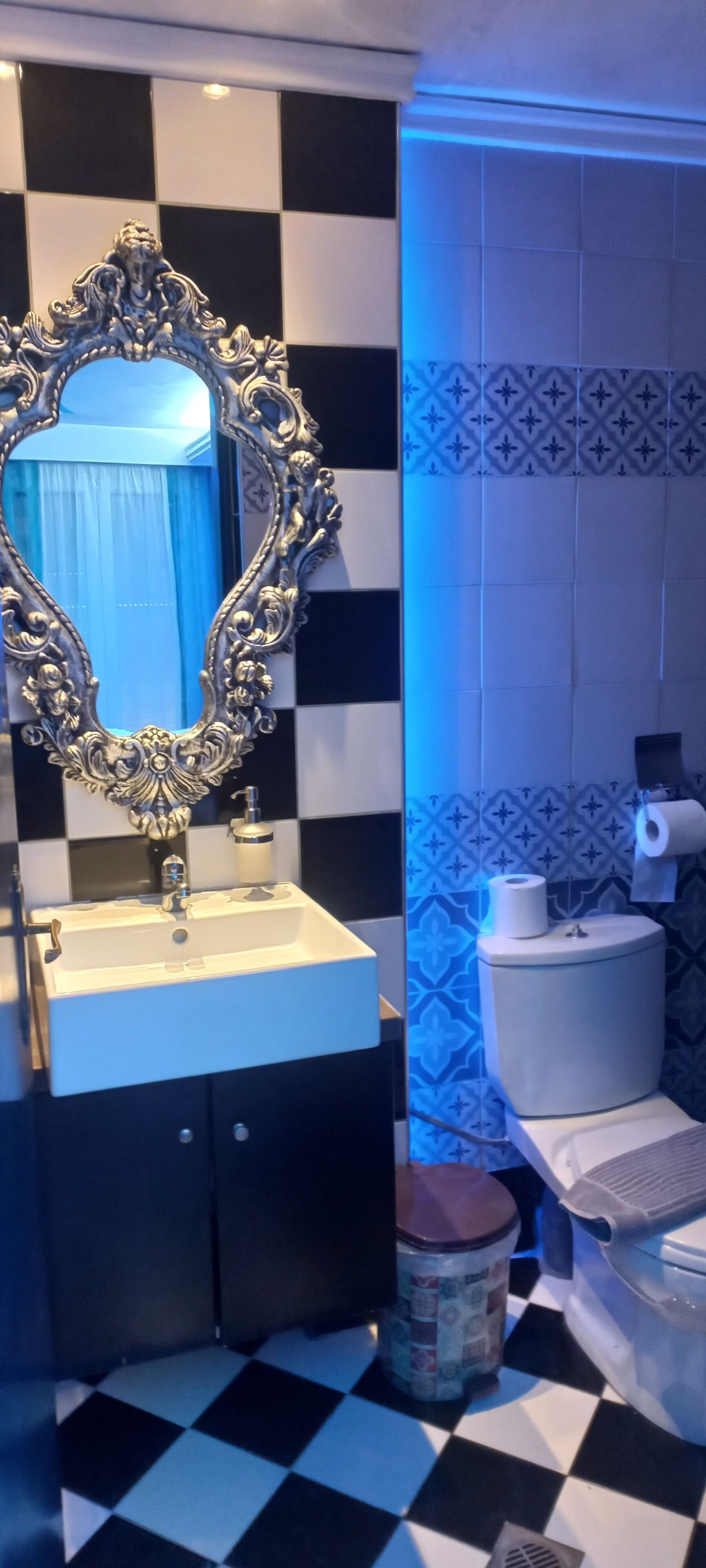 A bathroom with a checkered floor, a sink with a decorative mirror, a toilet, and a trash bin.