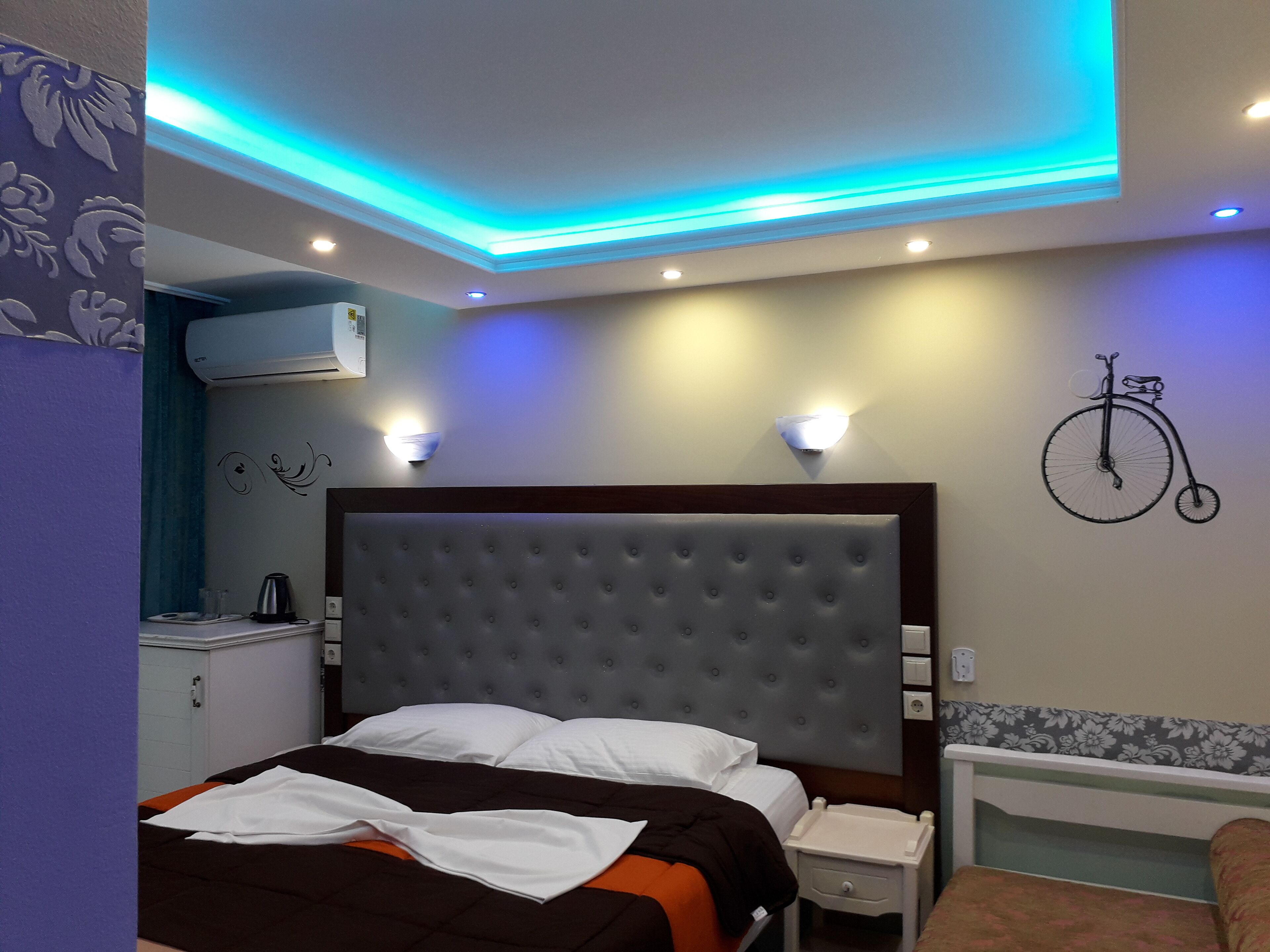 A bedroom with a tufted headboard, a bedside table, a wall-mounted bicycle decoration, and a ceiling with blue LED lighting.