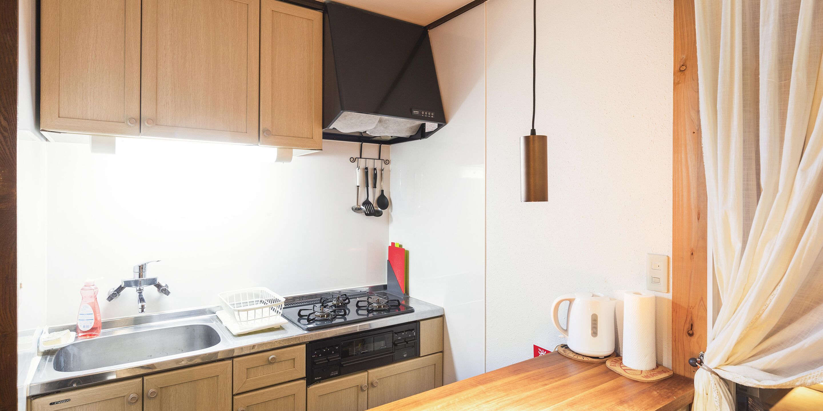 Cocina compacta con gabinetes de madera, un lavamanos de acero inoxidable, una estufa a gas y una campana extractora negra.