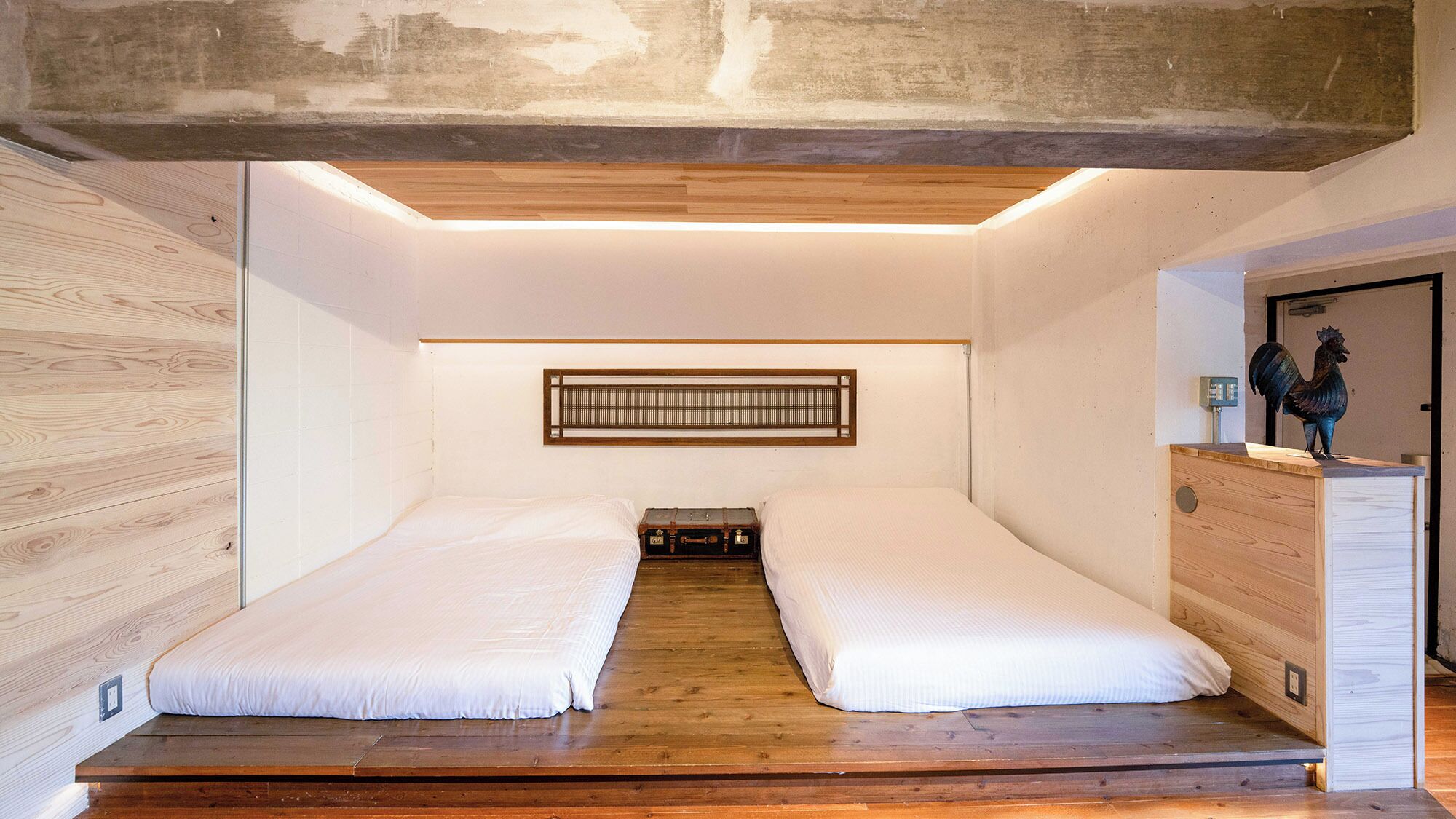 Une chambre avec deux lits, un plancher en bois, un radiateur fixé au mur et une commode en bois surmontée d’une statuette de coq.