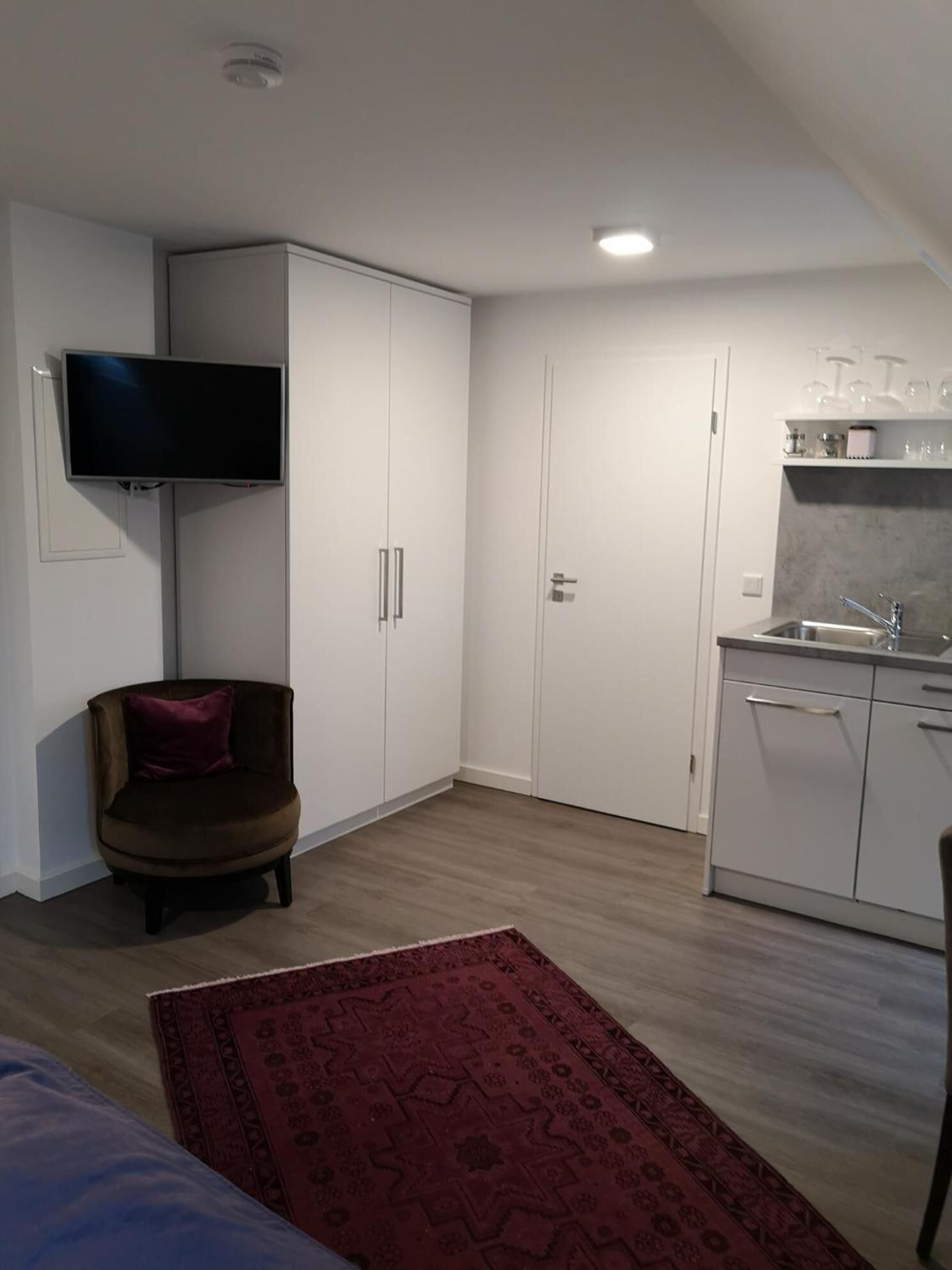 A compact room with a kitchenette, a flat-screen TV mounted on the wall, a brown chair with a red cushion, and a patterned rug on the floor.