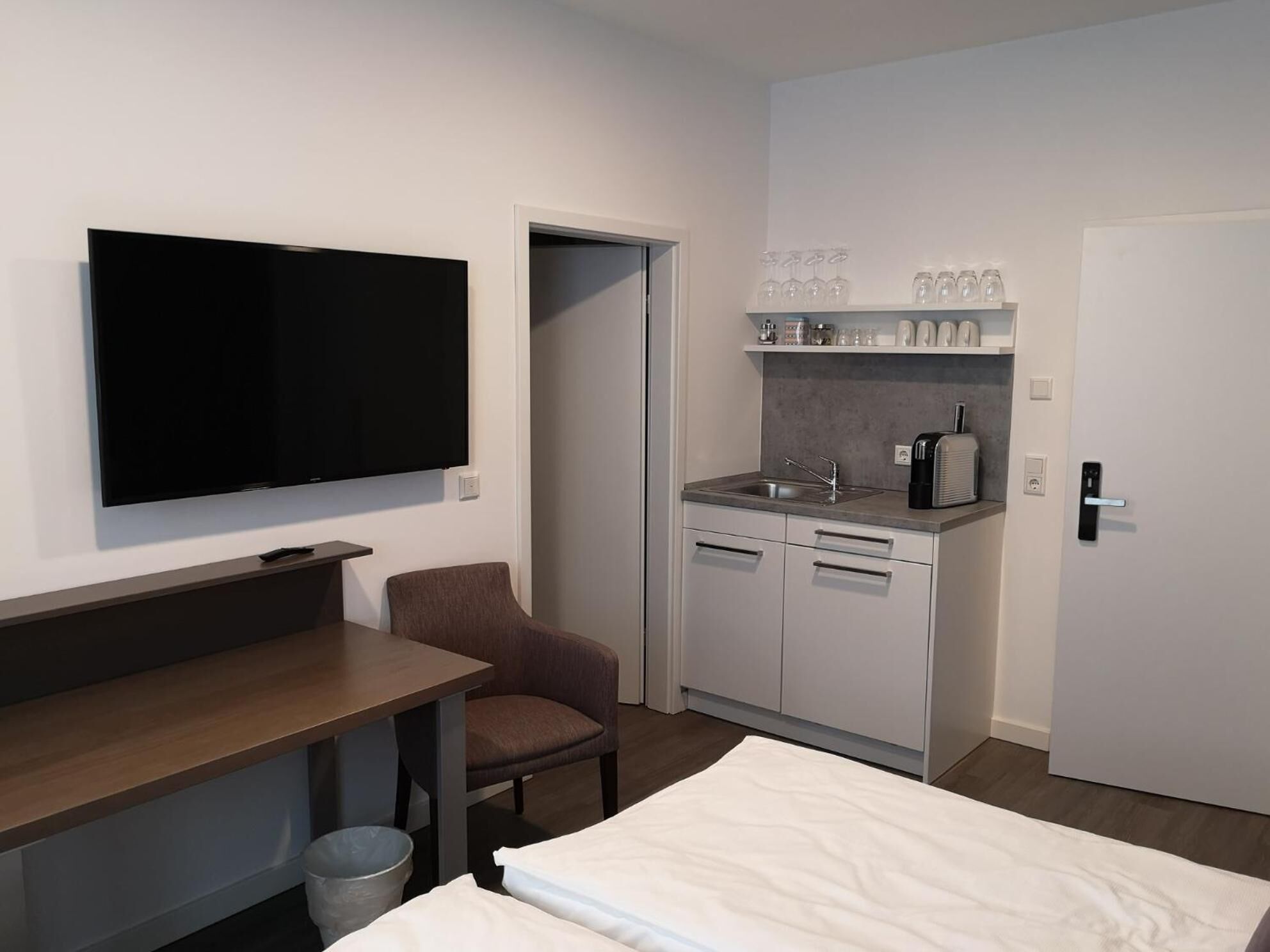 Habitación de hotel con televisión de pantalla plana, un escritorio pequeño, una silla, una cama y una kitchenette con un lavabo.