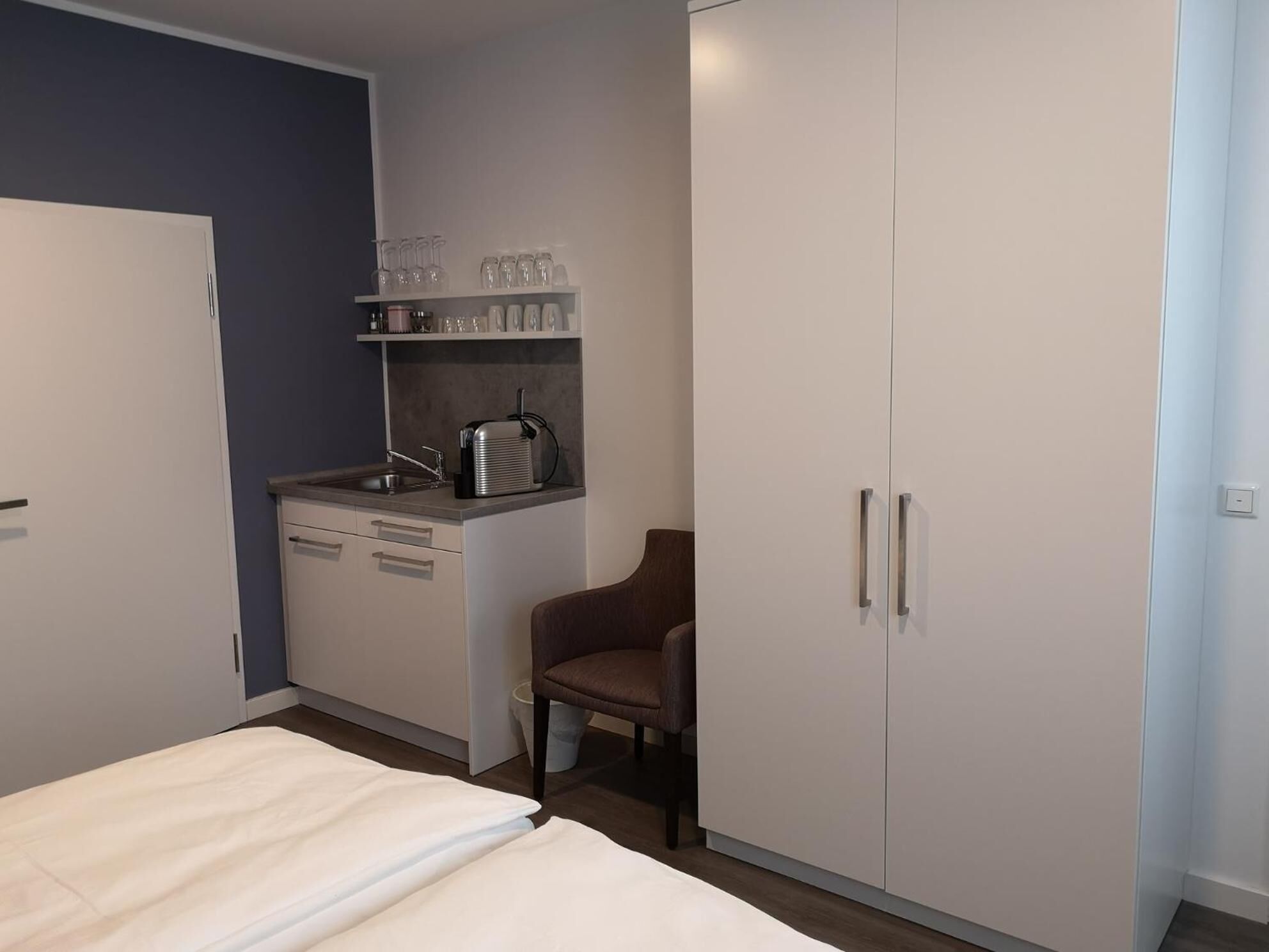 Un dormitorio con cama, una silla, armario, pequeña kitchenette y una estantería con vasos.