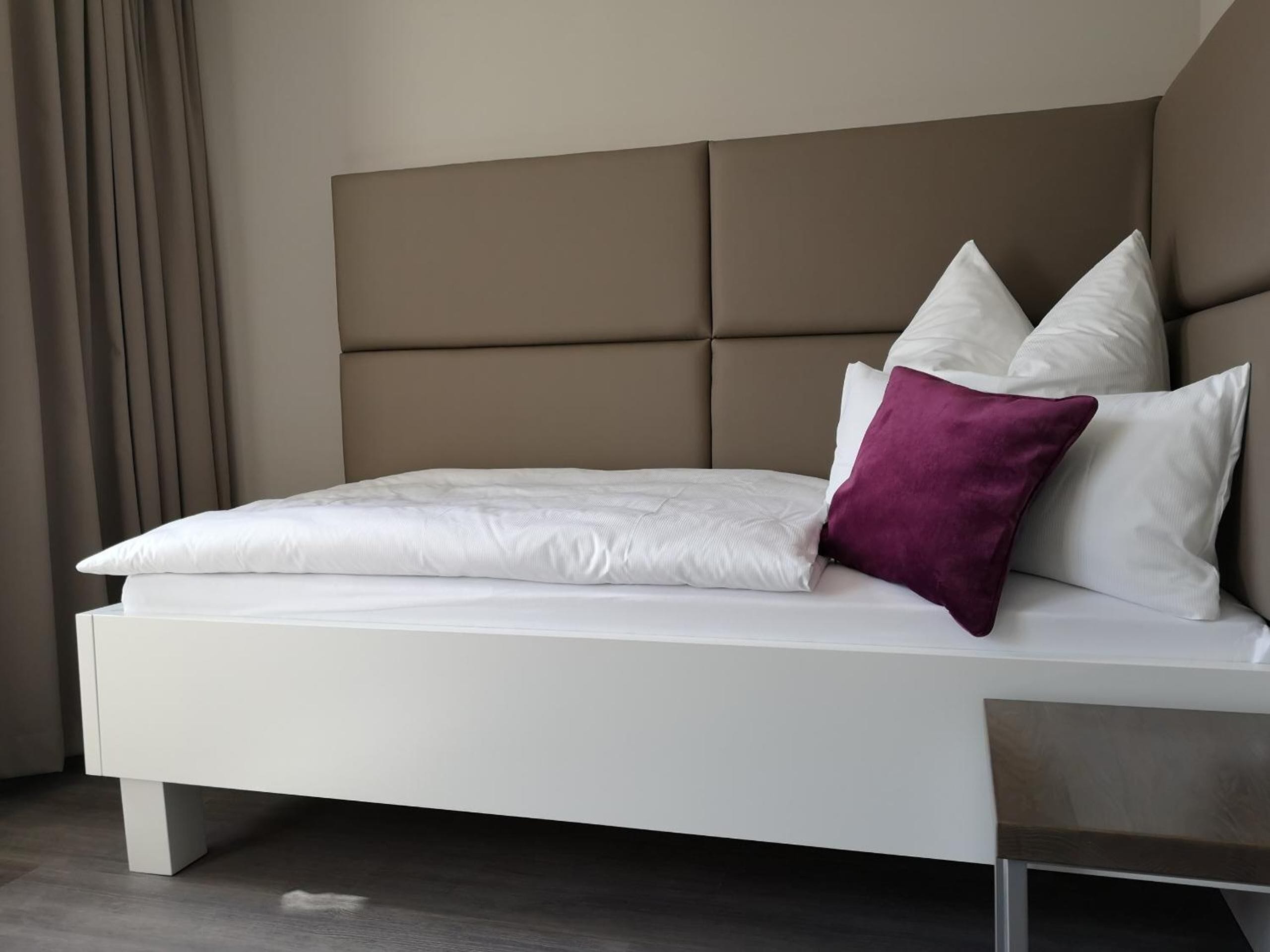 Un lit moderne doté d’une tête de lit rembourrée, de draps blancs et d’un coussin décoratif violet.