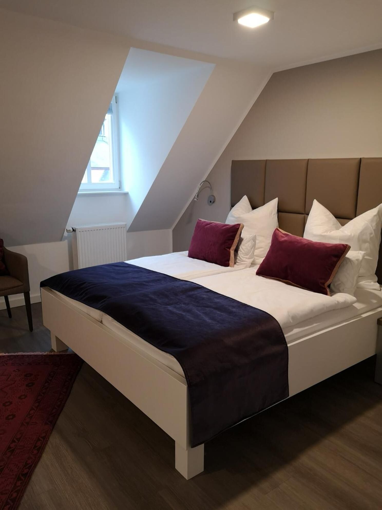 Een slaapkamer met een schuine kap, een bed met witte en bordeauxrode kussens, een donkerblauwe dekbedovertrek en een rood tapijt.