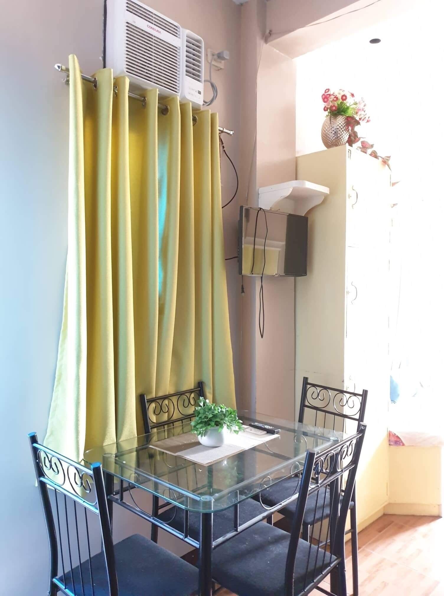 Un pequeño comedor con una mesa de vidrio y sillas de metal negro, una cortina amarilla y un aire acondicionado instalado en la pared.