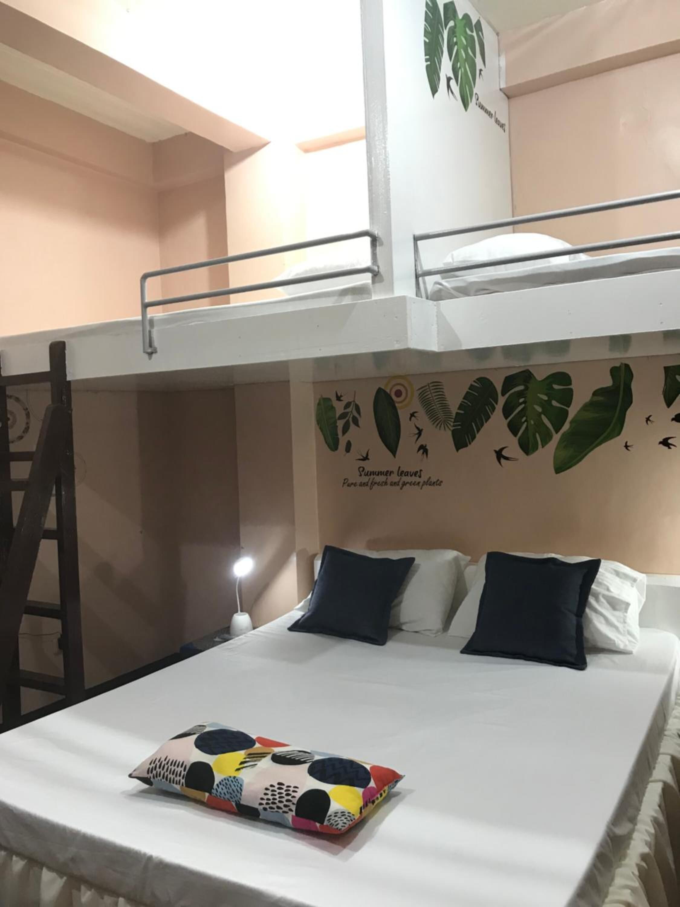 Une chambre avec un lit superposé, une échelle, une fresque murale représentant des feuilles et un lit agrémenté d’un coussin coloré.
