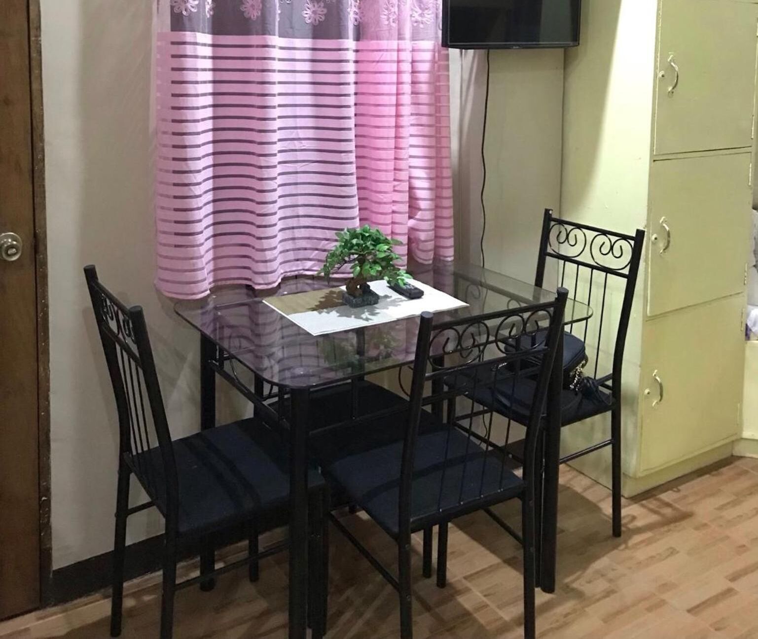 Un pequeño comedor con una mesa de vidrio y cuatro sillas negras, una cortina a rayas rosas, una pequeña planta en maceta y un televisor montado en la pared.