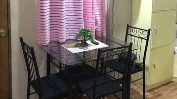 Un pequeño comedor con una mesa de vidrio y cuatro sillas negras, una cortina a rayas rosas, una pequeña planta en maceta y un televisor montado en la pared.