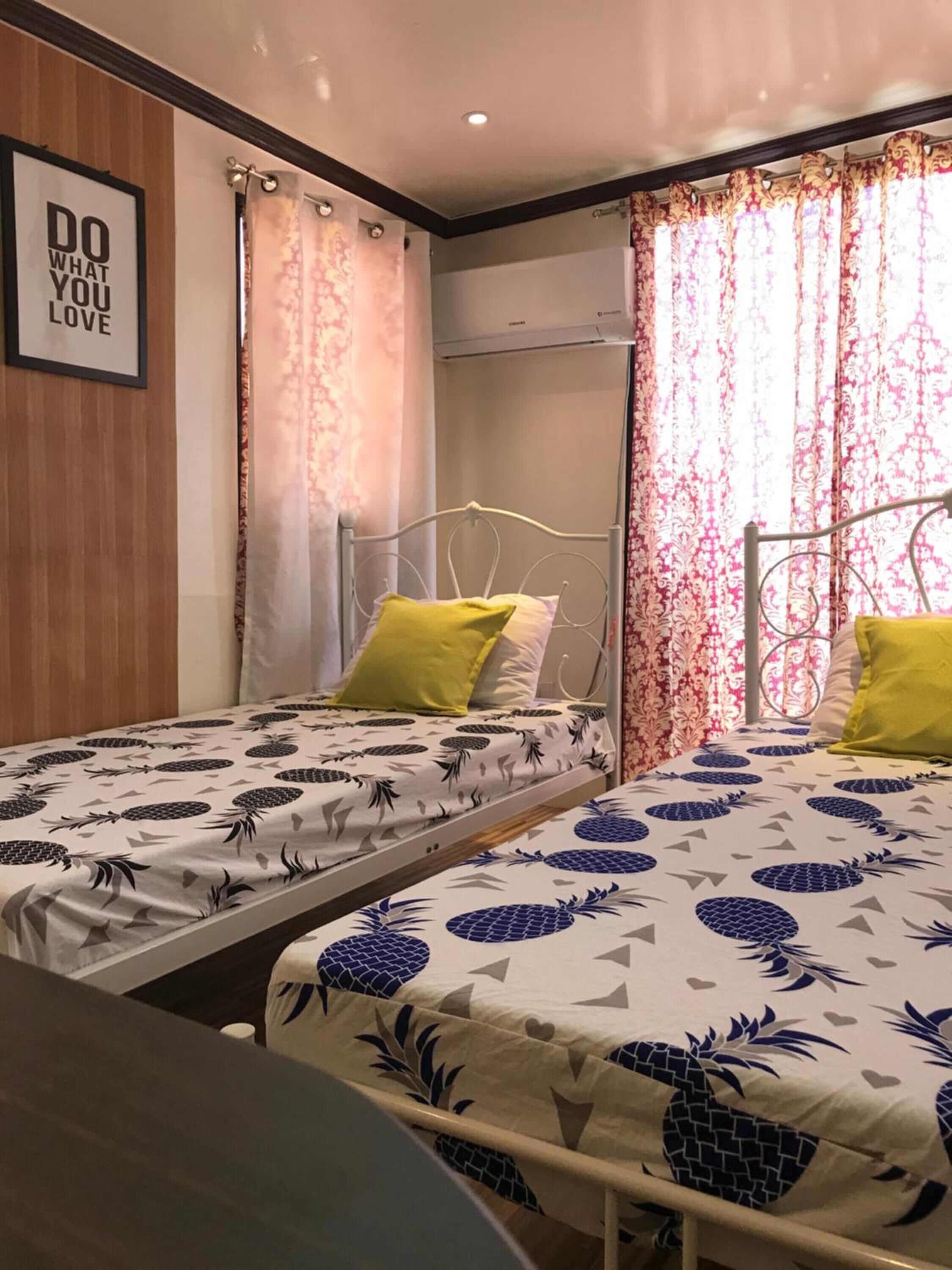 A room with two single beds, each with a pineapple-patterned bedspread. There is a framed motivational quote on the wall, and the room has a wooden panel design on one side.