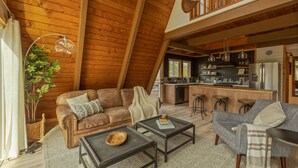 A cozy loft-style living room with a high ceiling, wooden beams, a kitchen area with bar stools, and a dining table. - Cozy A Frame Cabin, Bass Lake, Ca-Acorn Falls  (Bass Lake)