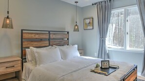 Ein Schlafzimmer mit Holz-Kopfteil, weißer Bettwäsche, einem Nachttisch und einer Wandlampe. Ein Fenster mit Vorhängen ist ebenfalls zu sehen.