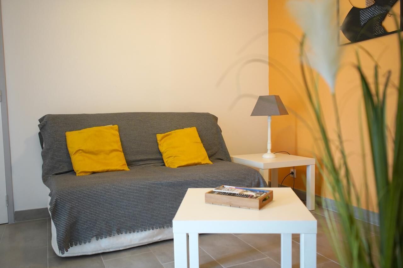 Una habitación con un sofá gris, dos cojines amarillos, una mesita auxiliar blanca con una lámpara y una pequeña mesa con un juego de mesa.