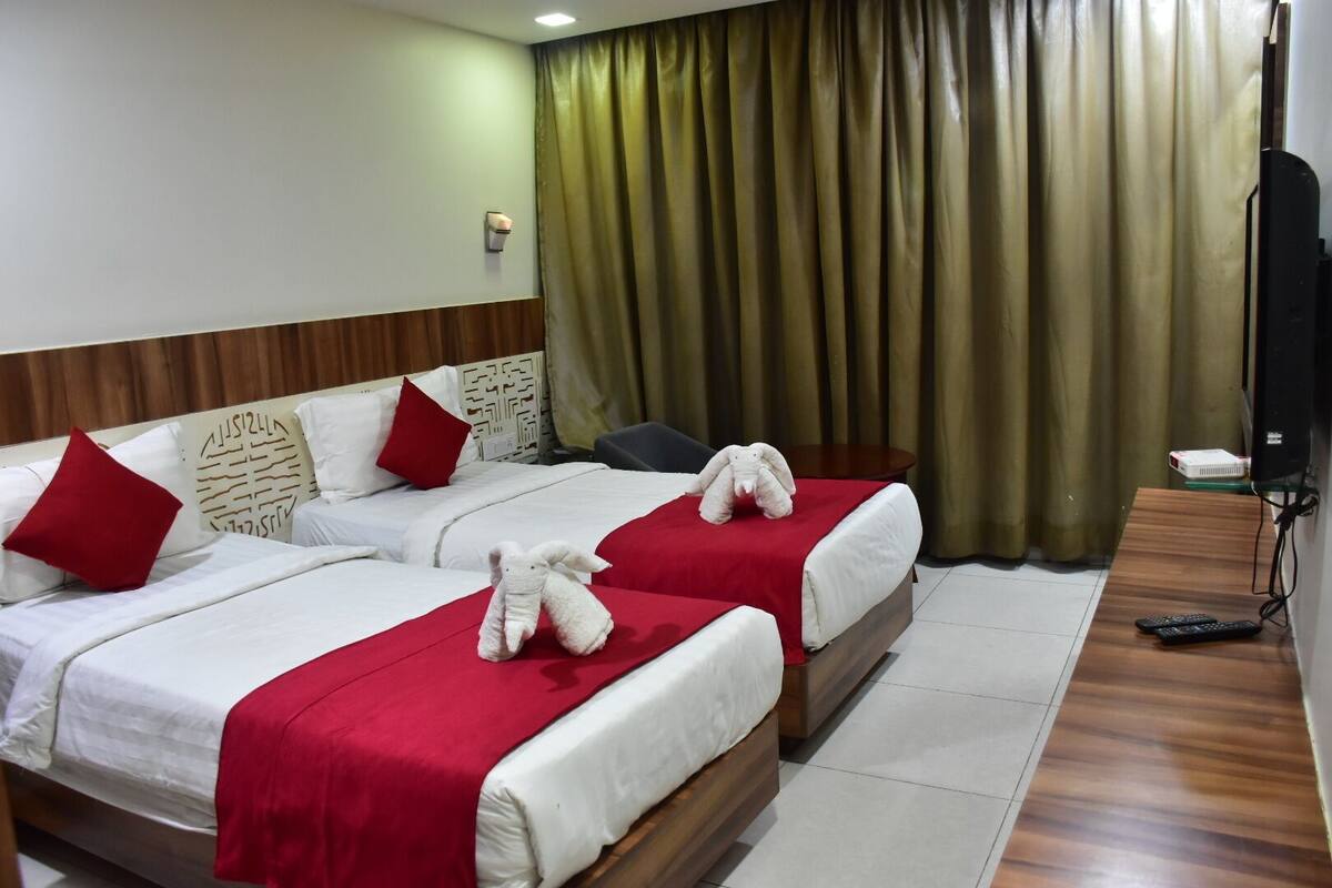 A hotel room with two beds, each with a red blanket and pillows, a wooden headboard, a small table, and a TV on the wall.
