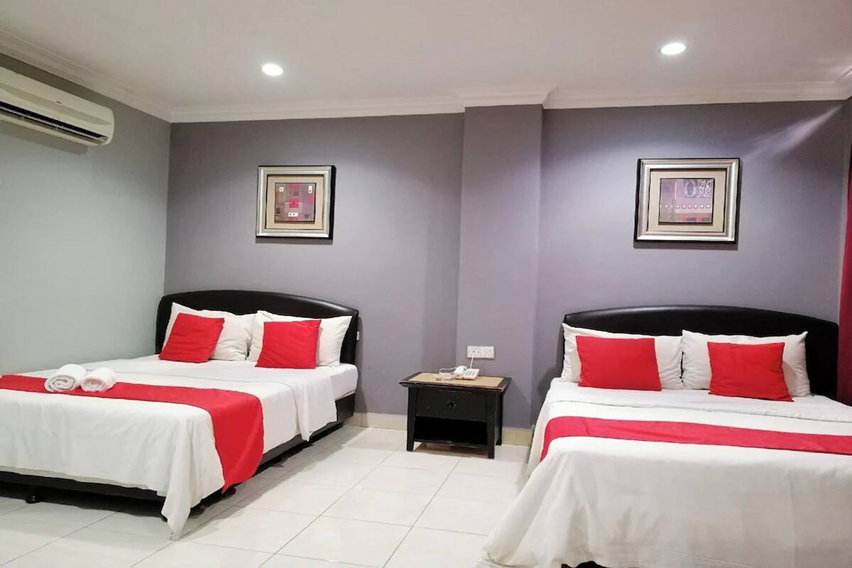 Quarto de hotel com duas camas, cada uma com roupa de cama branca e almofadas vermelhas, uma pequena mesa de cabeceira com um telefone e um aparelho de ar condicionado na parede.