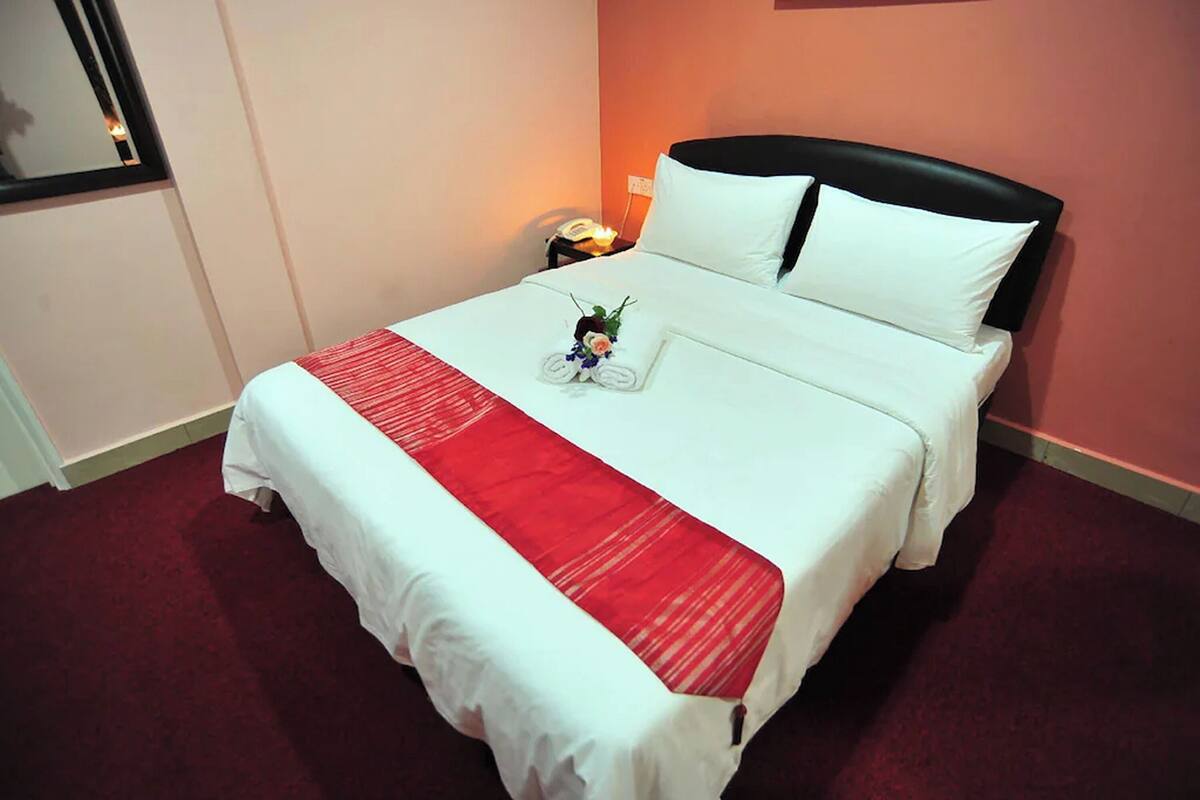 Cama bem arrumada com lençóis brancos, um corredor vermelho decorativo e um pequeno arranjo floral.