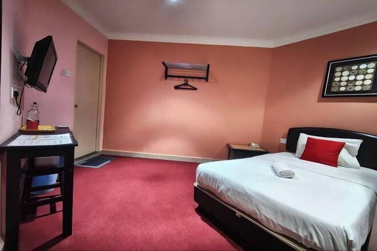 Quarto de hotel com uma cama, uma mesa com papéis, um cabideiro e uma televisão fixada na parede.