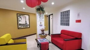 Un área de recepción con un sofá amarillo, un sofá rojo, una pequeña mesa con mantel rojo, un cuadro enmarco de la Torre Eiffel y una planta decorativa.