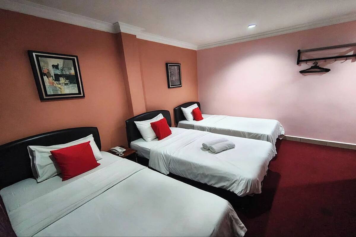 Quarto de hotel com três camas, cada uma com roupa de cama branca e almofadas vermelhas, um telefone ao lado esquerdo e um quadro na parede.