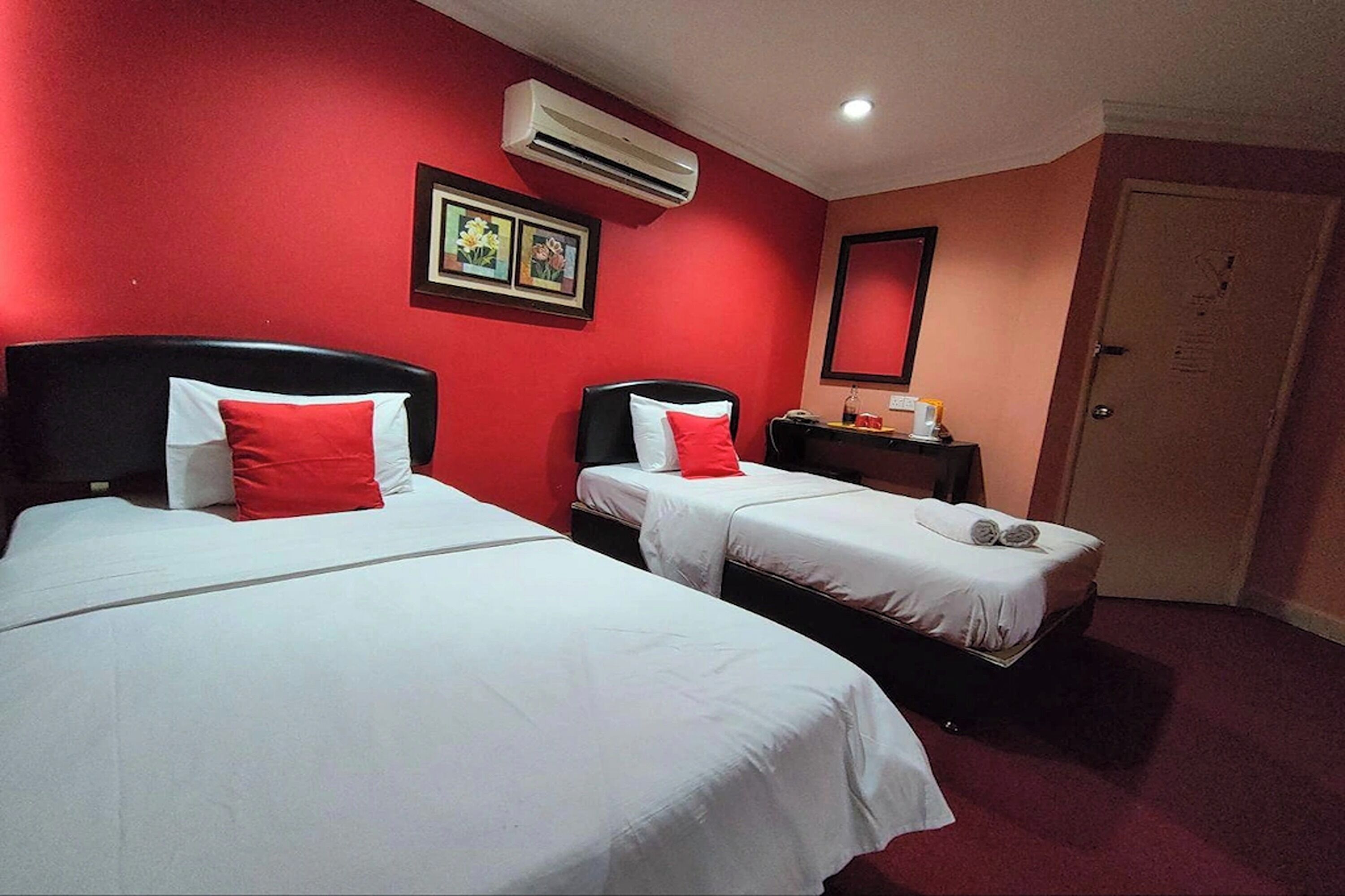 Two beds with red pillows, a bedside table with a lamp, and a wall-mounted air conditioner.