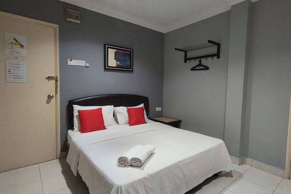 Quarto de hotel com cama, mesa de cabeceira, porta e cabideiro fixado na parede.