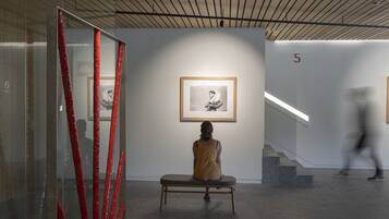 A modern art gallery with a minimalist design, featuring a glass partition with red accents, a bench, and framed artwork on the walls.