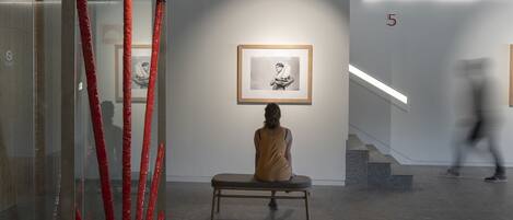 A modern art gallery with a minimalist design, featuring a glass partition with red accents, a bench, and framed artwork on the walls.