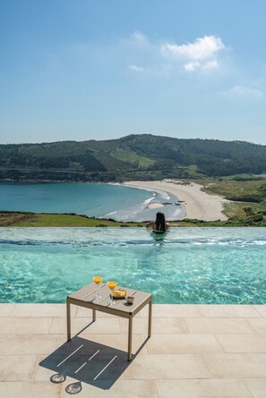 An infinity pool with a table and drinks, overlooking a beach and forested hills. - Parador Costa da Morte (Muxía)