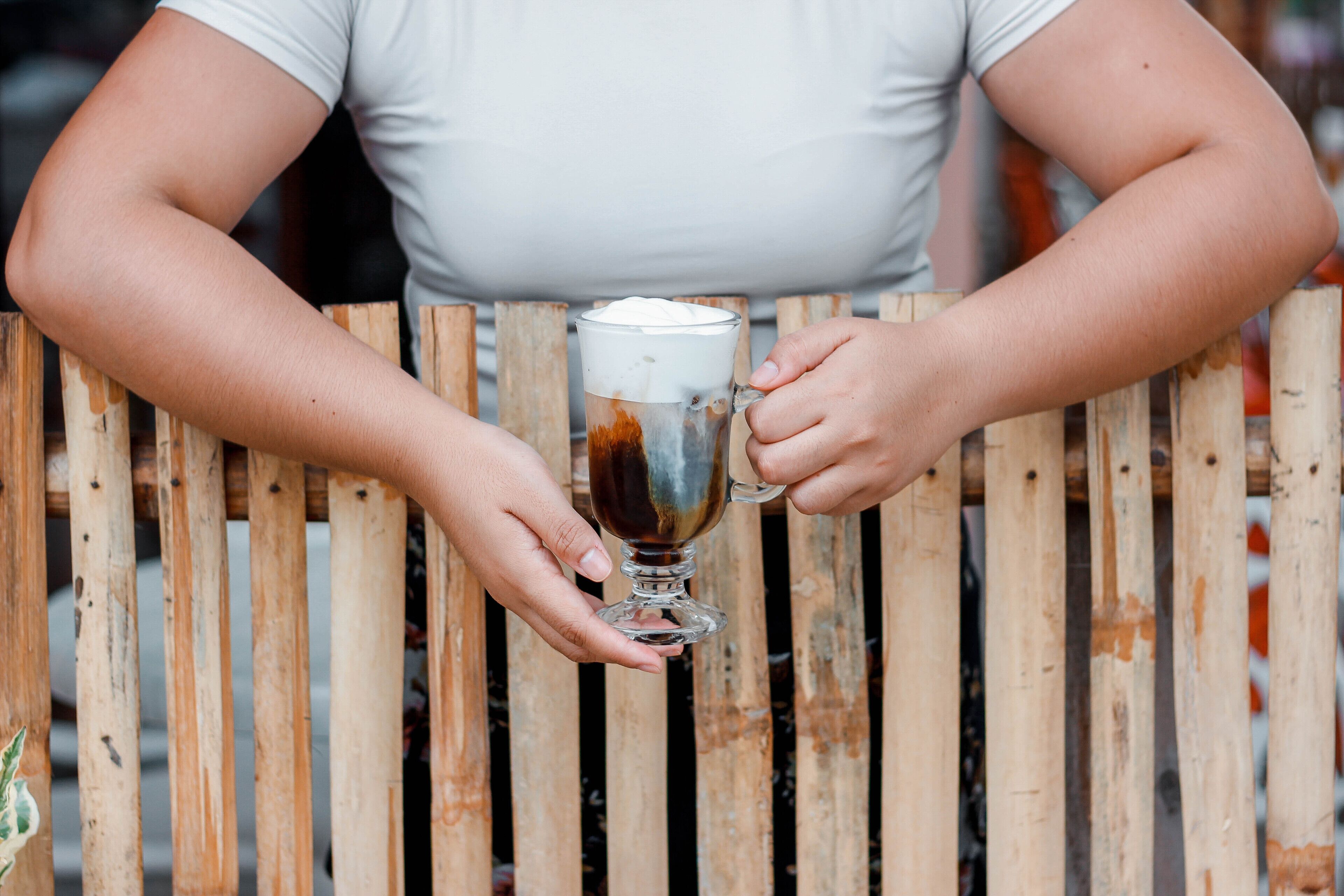 A person holding a glass mug with a creamy top, seated at a wooden table.