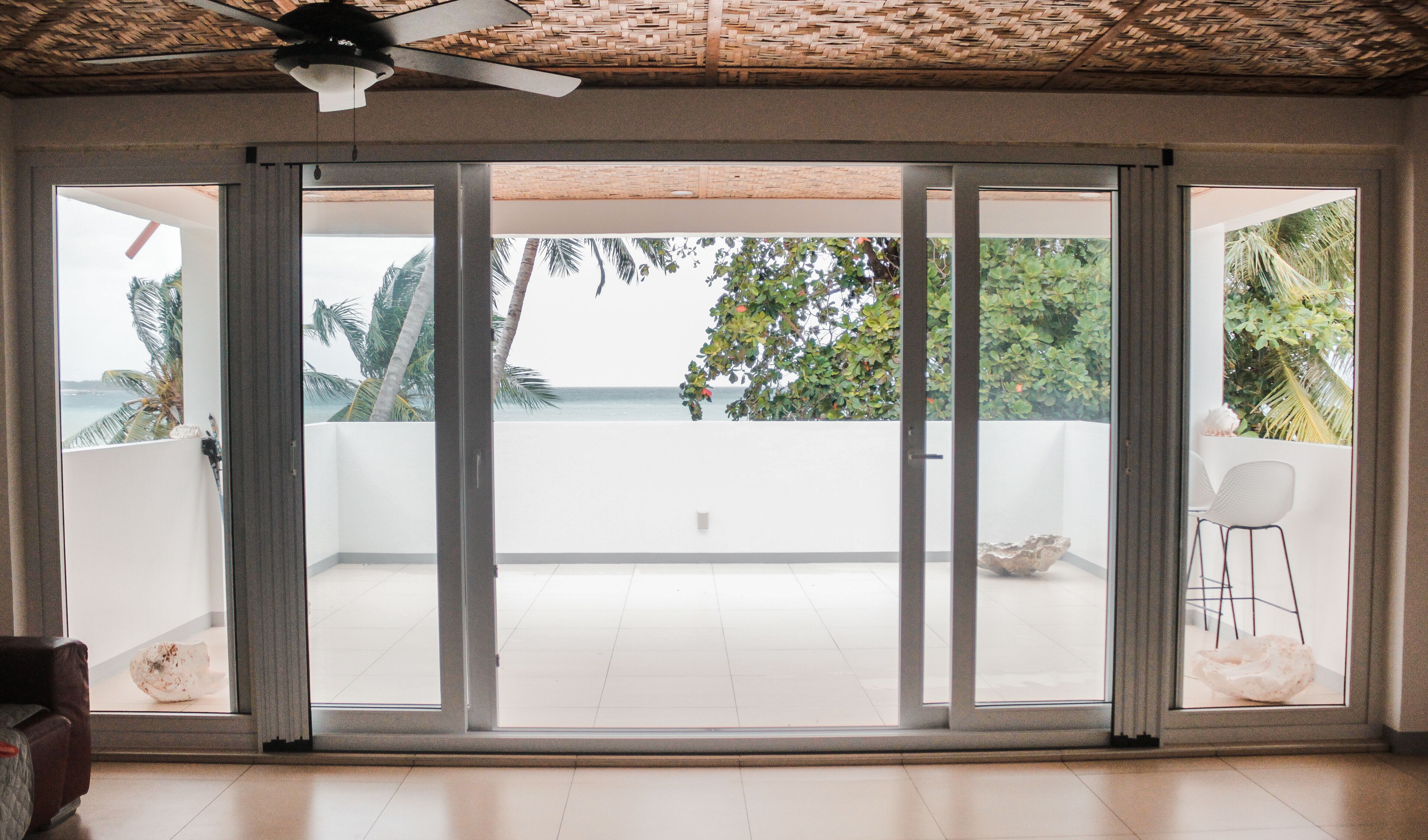 A room with a large sliding glass door leading to a balcony with a view of the ocean and palm trees.