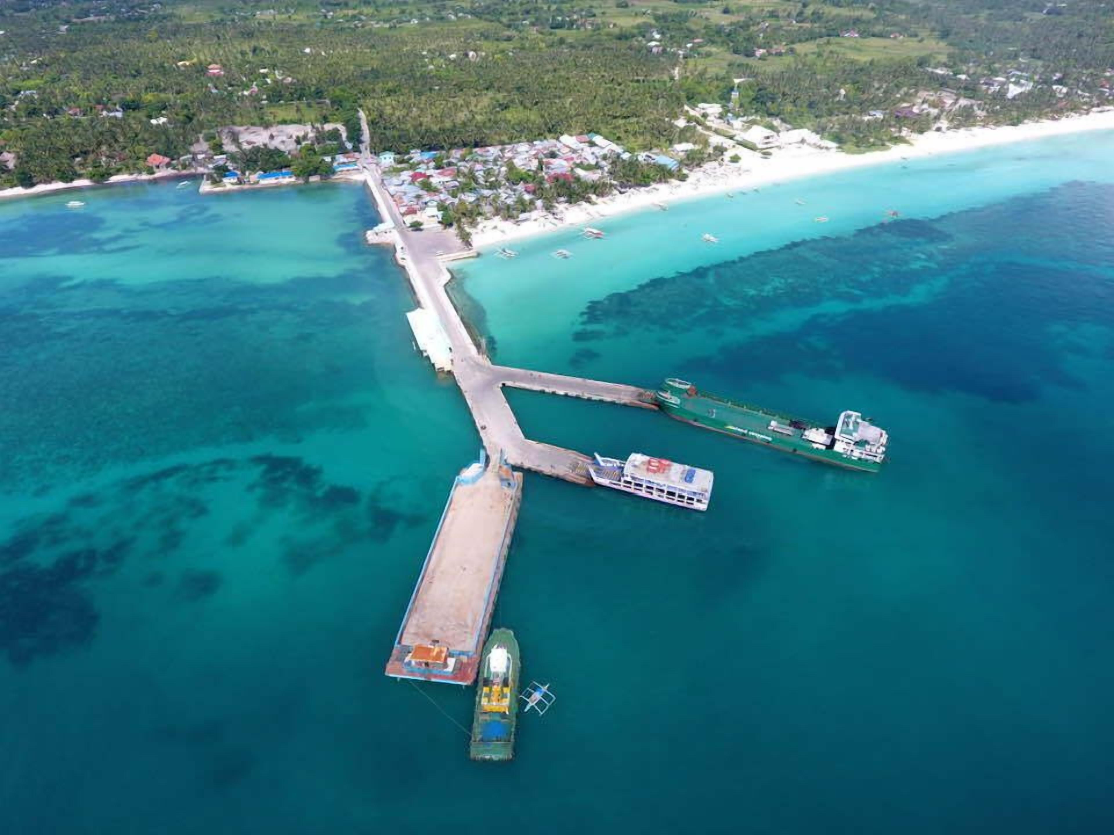 Vista aérea de una zona costera con un muelle que se adentra en aguas turquesas, un barco pequeño amarrado al muelle, y una playa con edificios dispersos al fondo.
