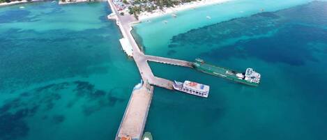 Vista aérea de una zona costera con un muelle que se adentra en aguas turquesas, un barco pequeño amarrado al muelle, y una playa con edificios dispersos al fondo.