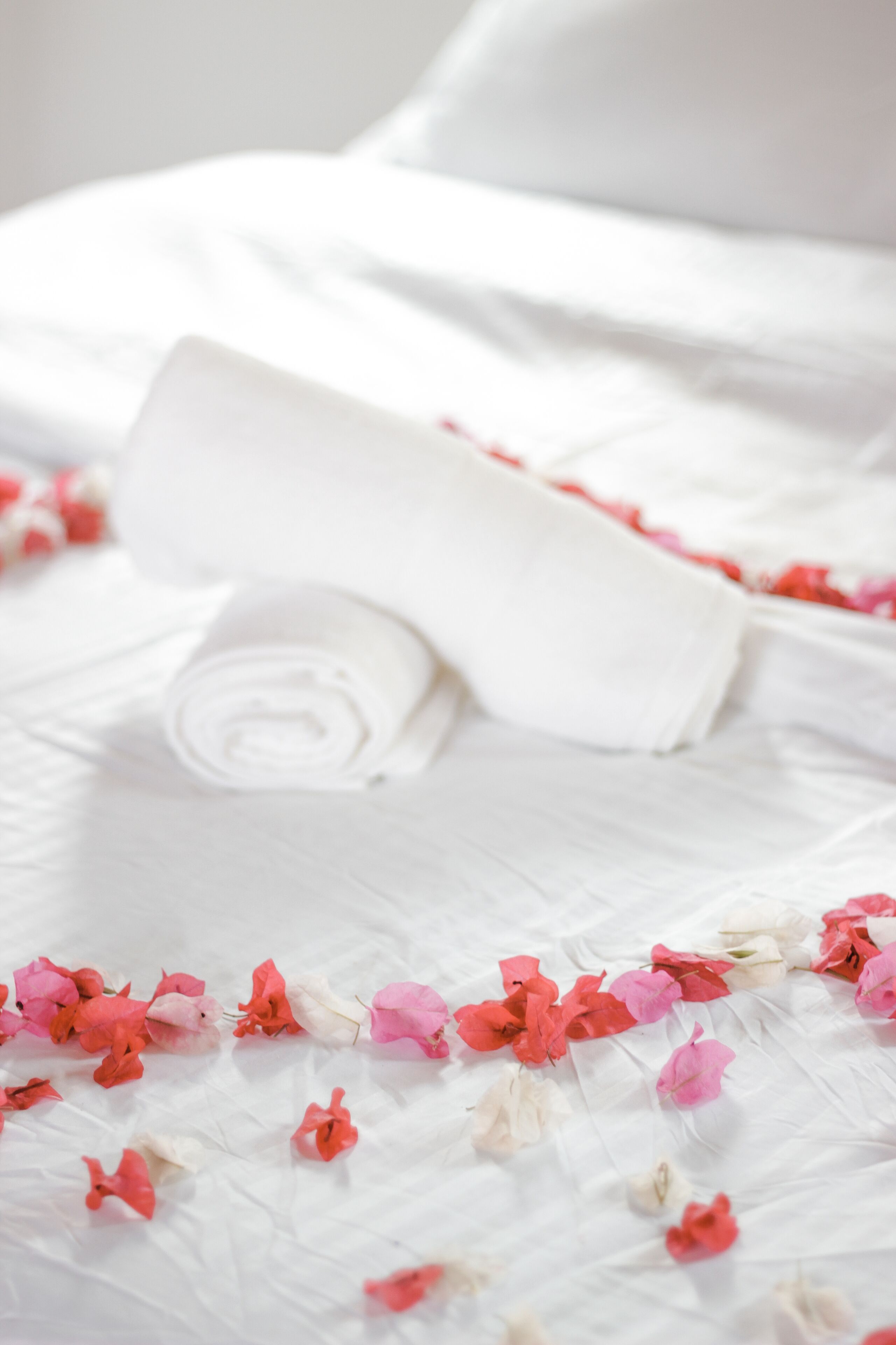 A neatly made bed with a rolled white towel and scattered pink and white flowers.