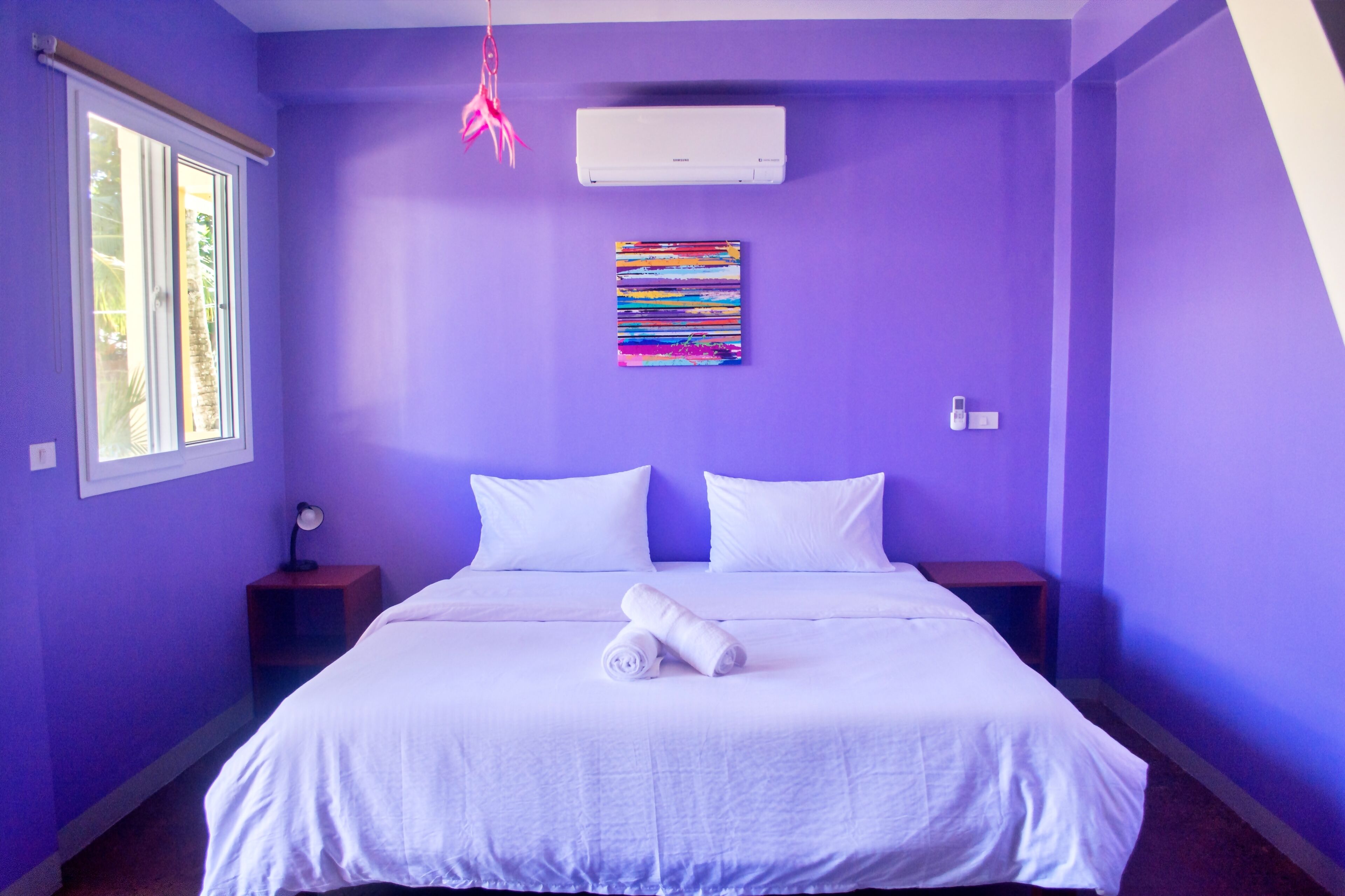 A room with a bed, white bedding, a colorful artwork on the wall, an air conditioner, and a window with a view.
