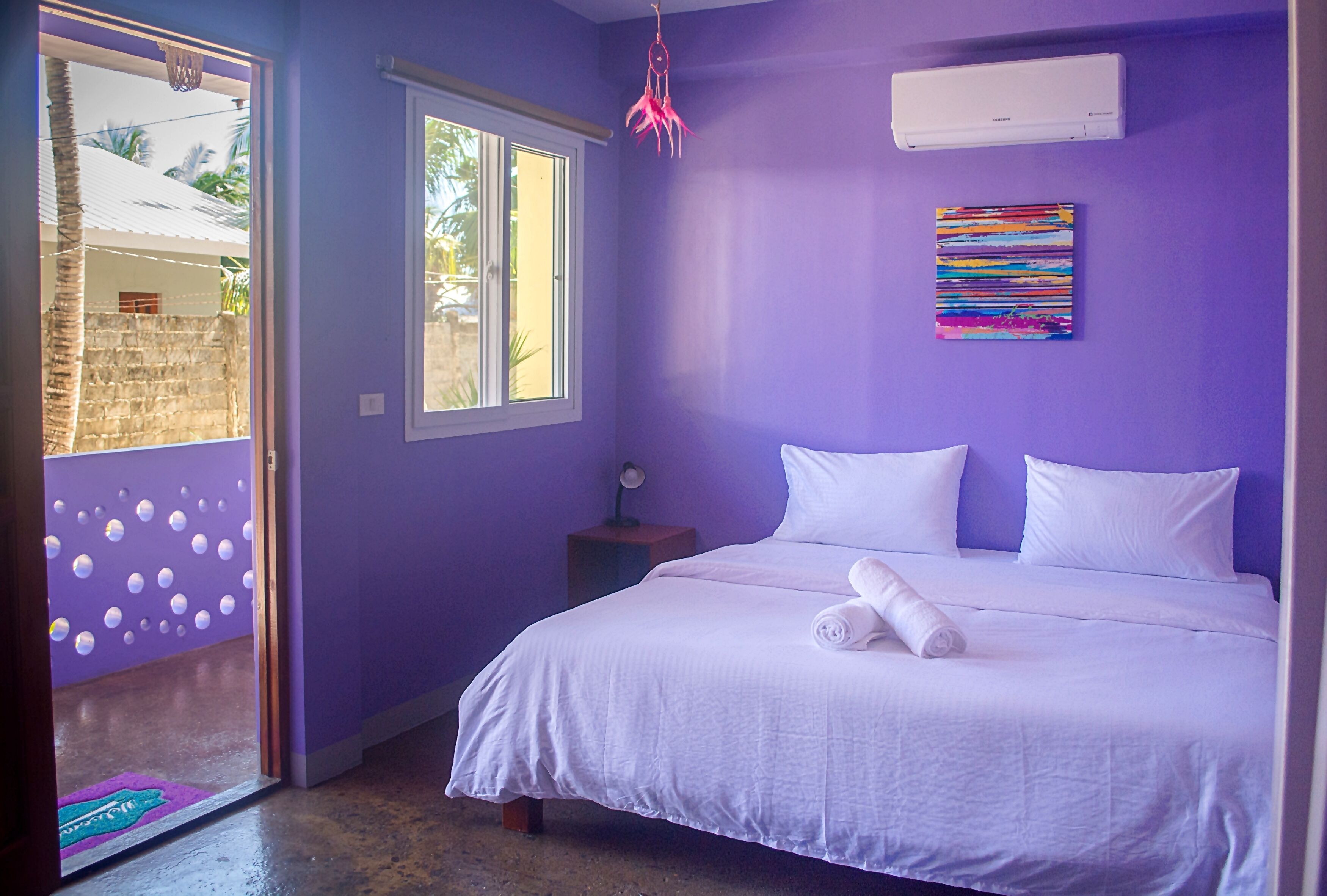 A bedroom with a bed, a window, a wall-mounted air conditioner, and a colorful abstract painting.
