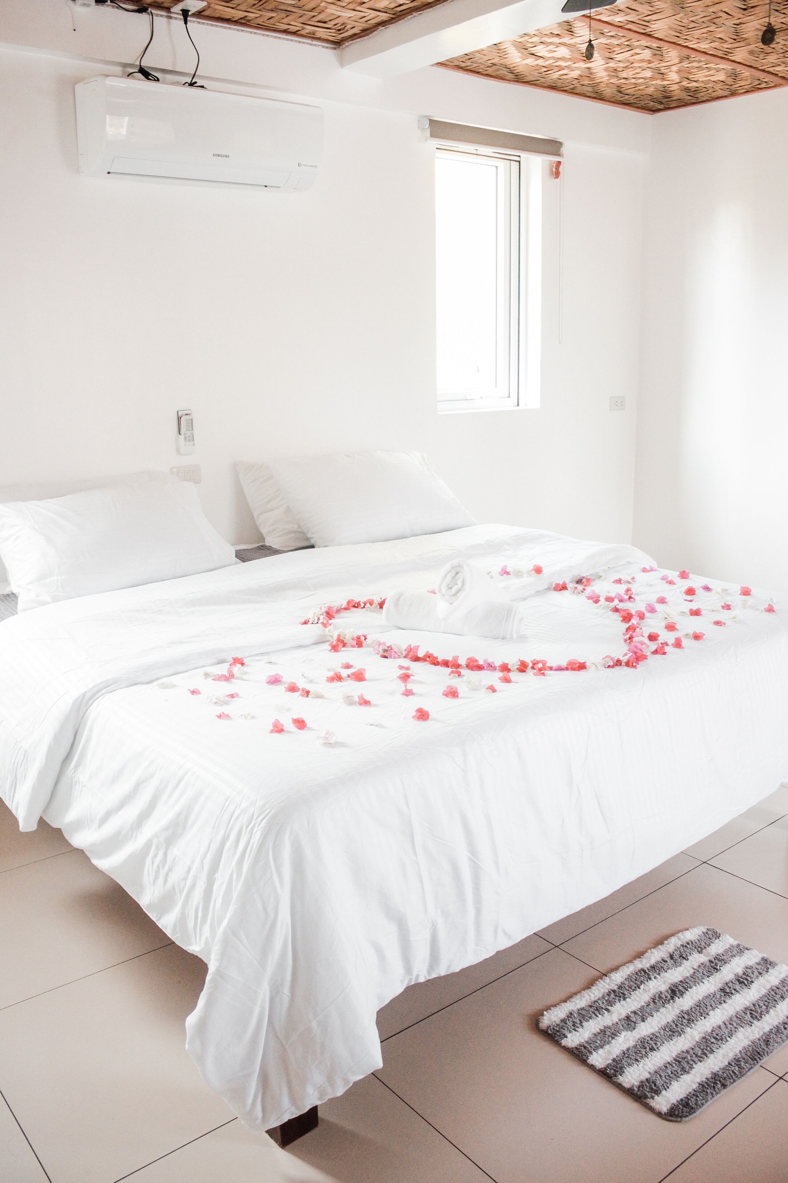 A white bed with rose petals, a striped rug, and a ceiling fan.